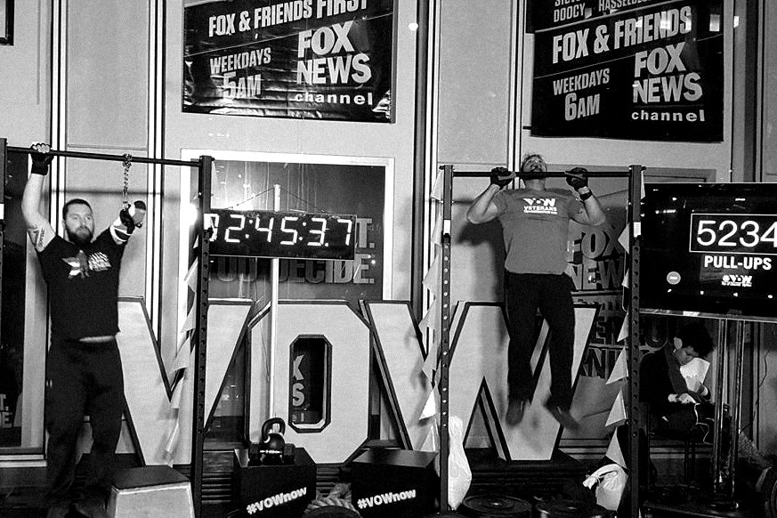 Guy mid pull-up on an outdoor rig in Times Square, a race clock behind him and a counter reading 5234 pull-ups.