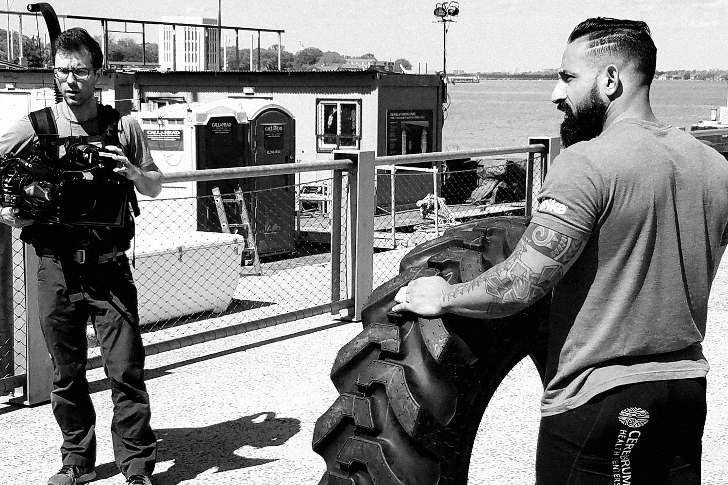 Guy resting a hand on a large tractor tire on a waterfront lot while a camera operator films him. Black and white.