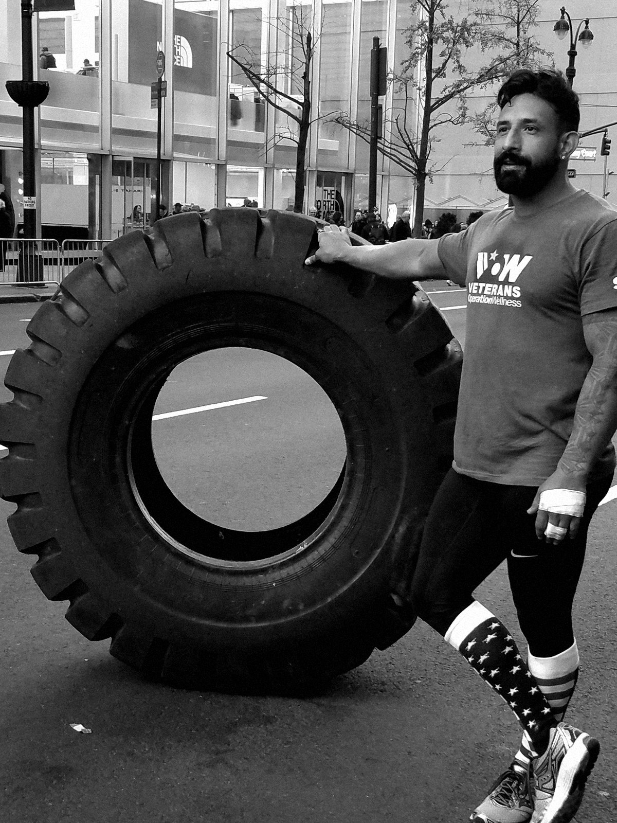 Guy in a Veterans Operation Wellness T-shirt, one hand on a large tractor tire on a city street. Black and white.